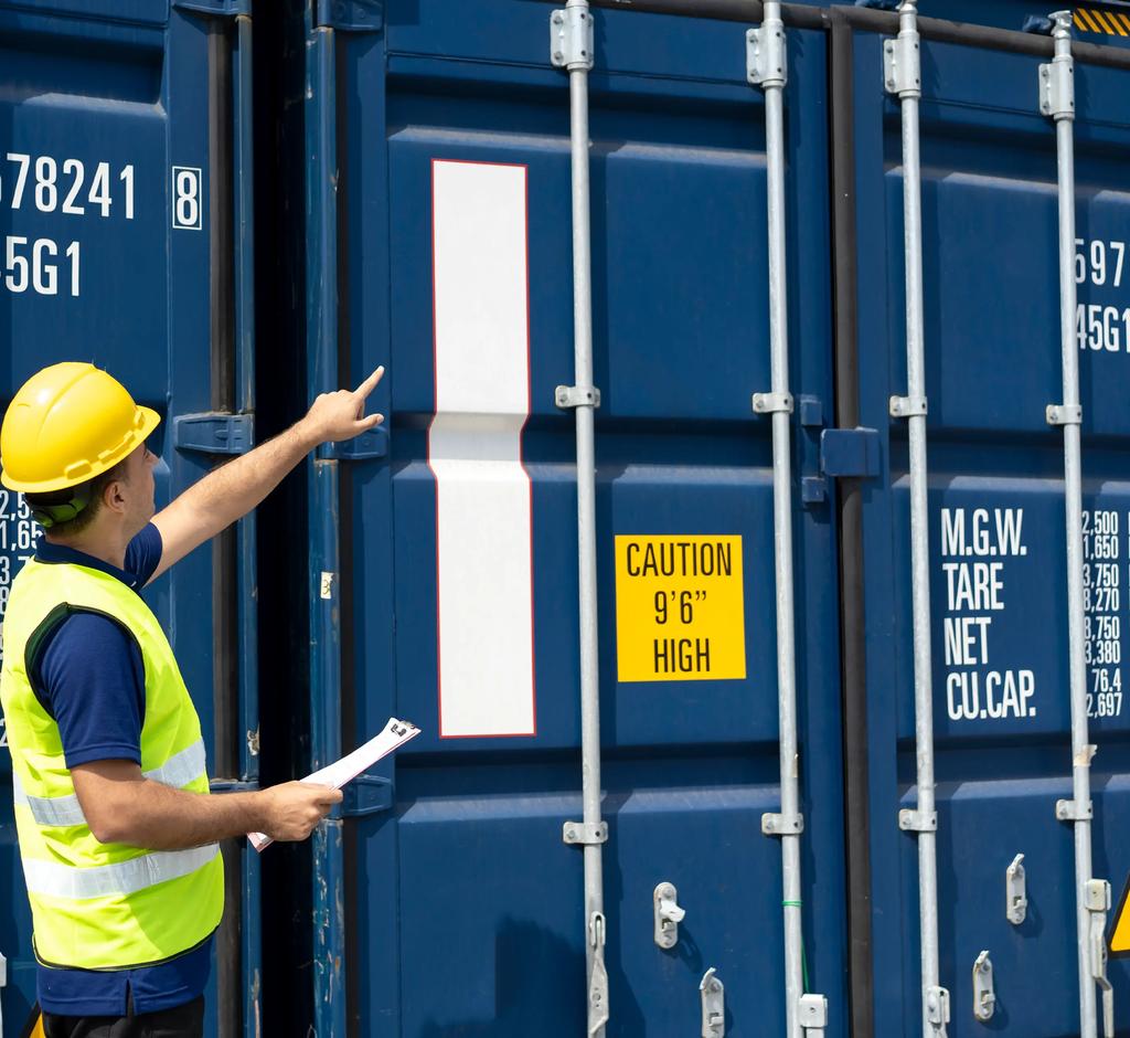 Photo of a worker in a hard hat and high-visibility vest holding a clipboard and pointing up at the markings on a blue shipping container.