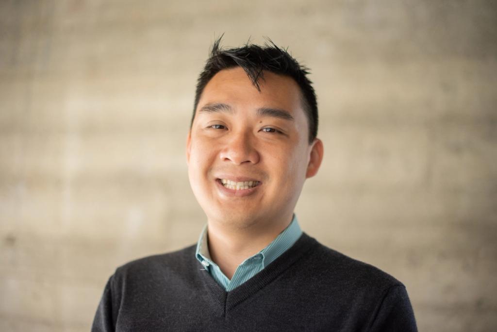 Headshot of a smiling man in a dark V-neck sweater over a striped shirt against a concrete wall