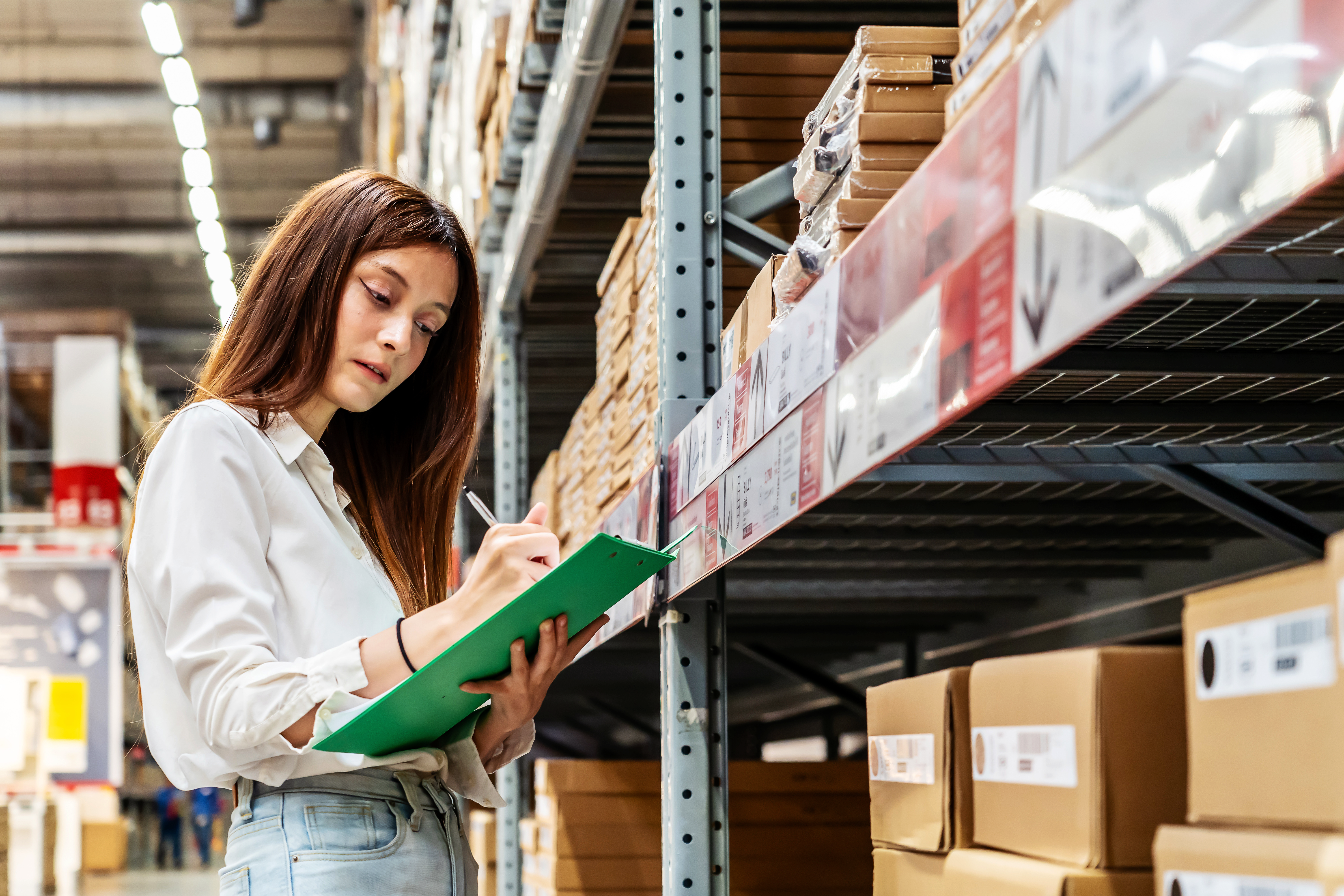 Woman in warehouse checking inventory list on clipboard while standing near storage shelves.