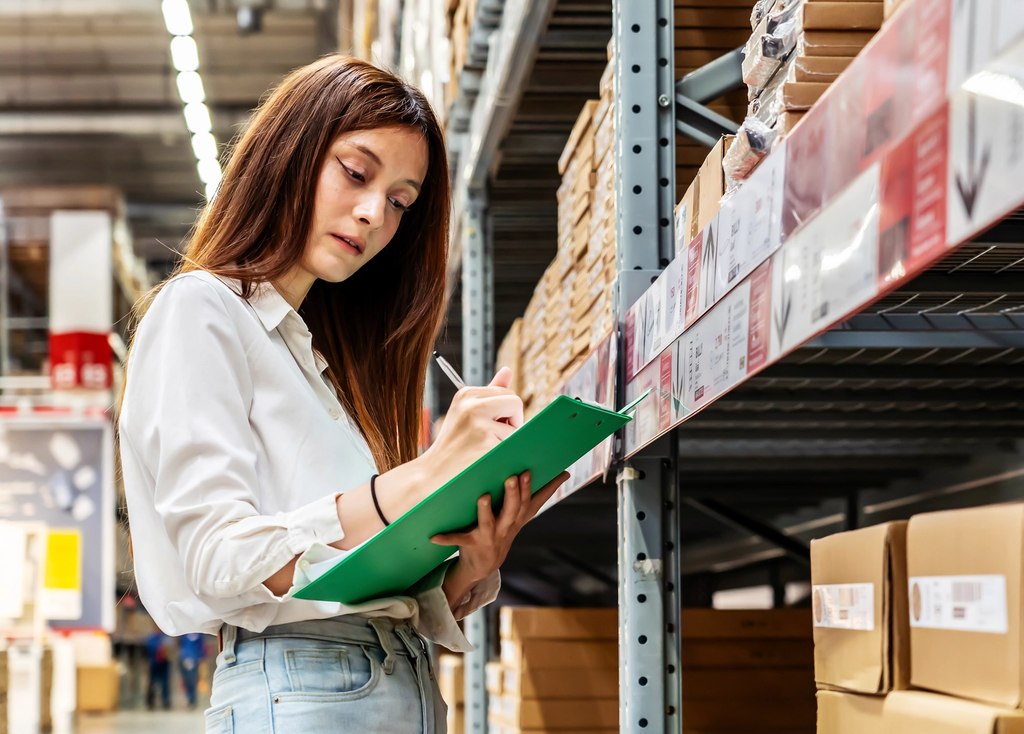 Woman in warehouse checking inventory list on clipboard while standing near storage shelves.