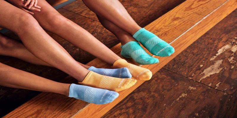 Three people's feet in colorful Bombas no-show socks on a wooden floor