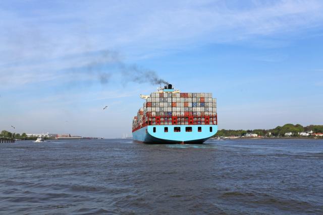 Blue container ship emitting dark smoke from its funnel as it sails along a river