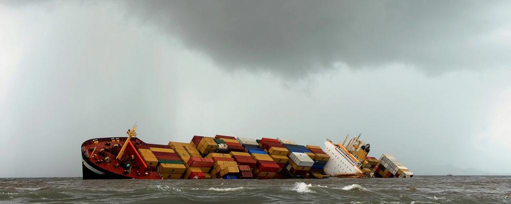 Container ship listing heavily to one side with stacked containers under stormy grey skies at sea