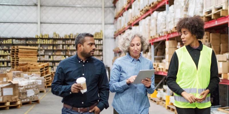 Three colleagues with a tablet walk through a warehouse; one wears a yellow hi-vis vest