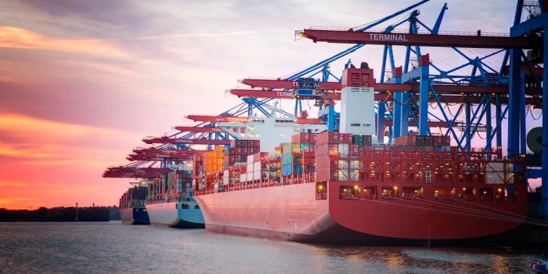 Container ships berthed at a terminal under cranes labeled "Terminal" at a pink-and-orange sunset.