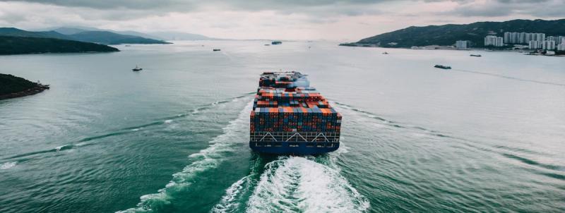 Aerial rear view of a loaded container ship sailing through a bay flanked by green hills
