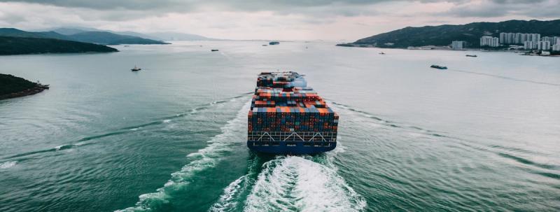 Aerial rear view of a loaded container ship sailing through a bay flanked by green hills