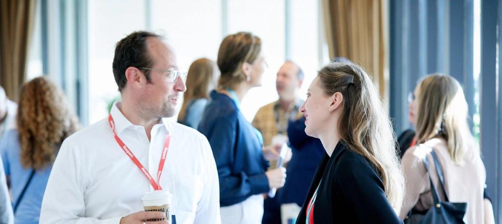 Two attendees with red lanyards talking, holding coffee cups, among a crowd at an indoor event