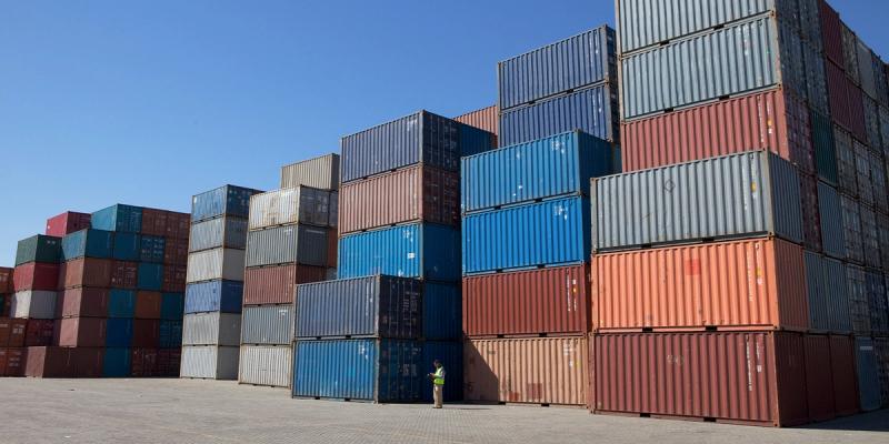 Stacks of shipping containers in a yard with a tiny hi-vis worker at the base for scale
