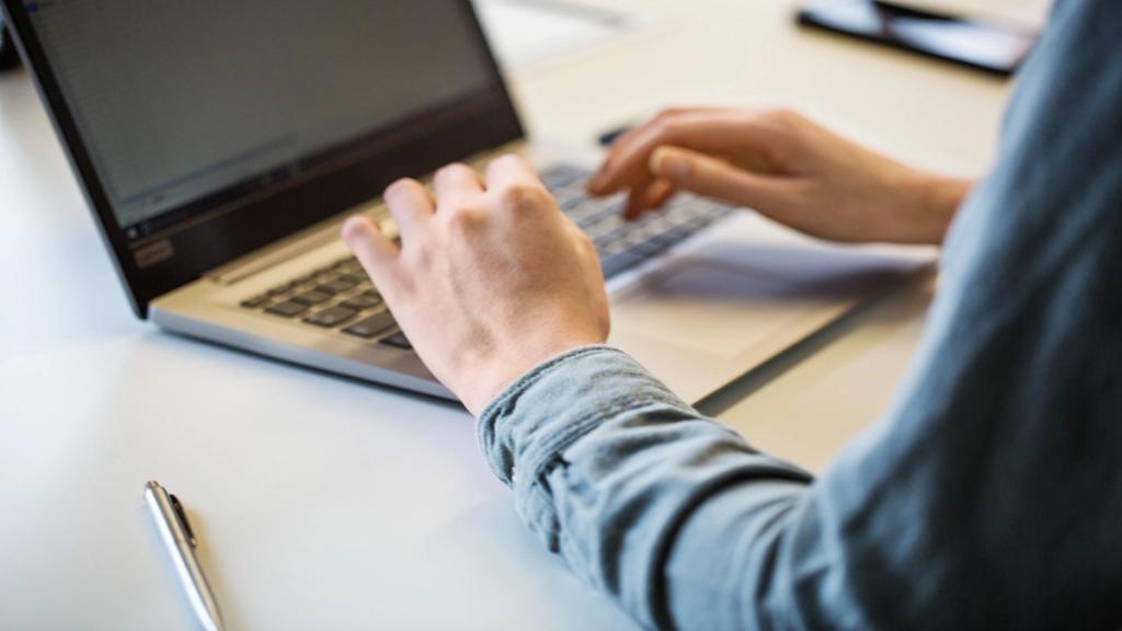Close-up of a person in a denim shirt typing on a laptop at a desk with a pen nearby