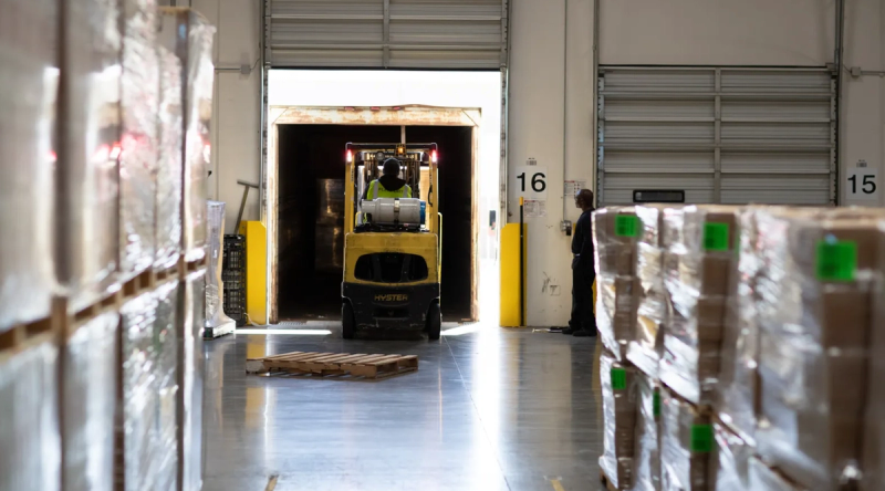 Forklift driving toward an open loading dock door in a warehouse, with stacked, shrink-wrapped pallets lining both sides of the aisle