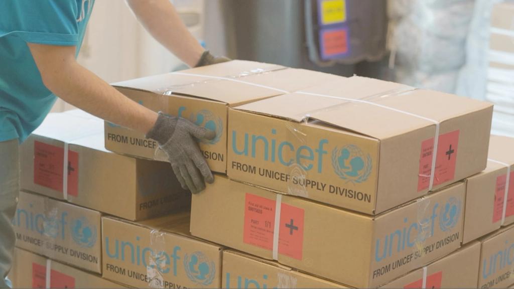 Worker in teal shirt and gloves stacking cardboard boxes labeled From UNICEF Supply Division with red first-aid kit labels