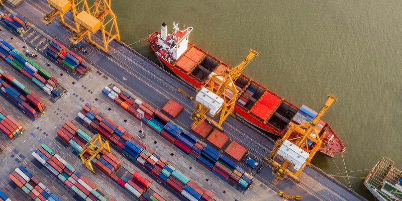 Aerial view of a red cargo ship docked at a container port with stacked shipping containers and gantry cranes