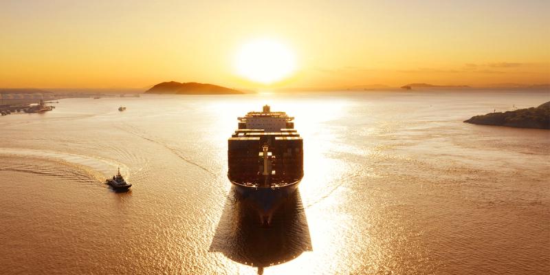 Aerial view of a container ship sailing toward a golden sunset with a tugboat alongside