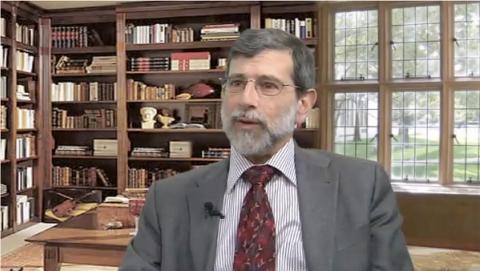 Bearded man in a gray suit and red tie speaking, seated in a library with wooden bookshelves