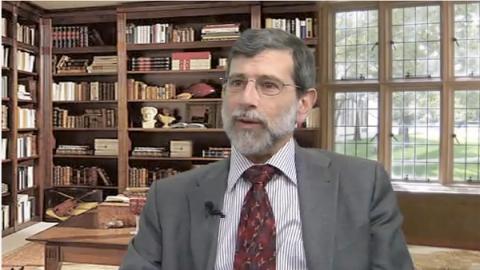 Bearded man in a gray suit and red tie speaking, seated in a library with wooden bookshelves
