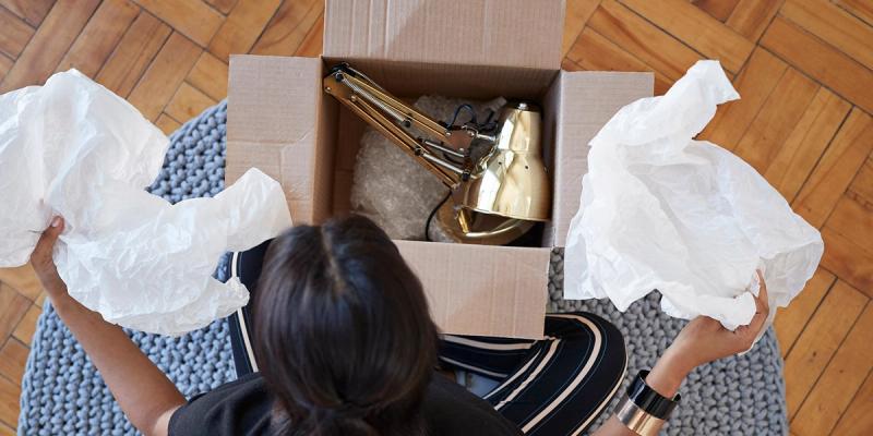 Overhead view of a woman unpacking a brass desk lamp from a cardboard box with white packing tissue