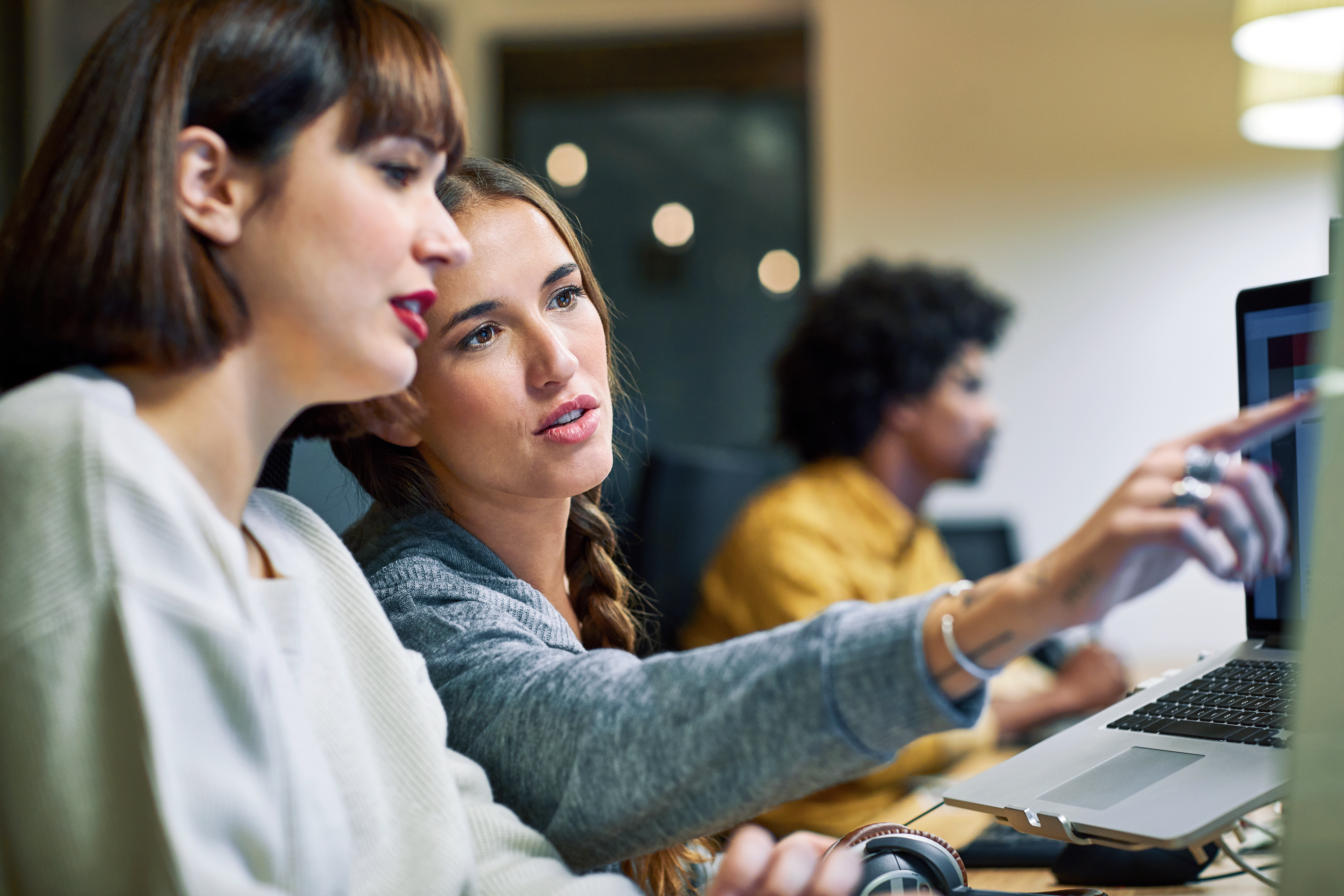 Two women collaborate at a laptop during a work meeting, pointing at the screen together.