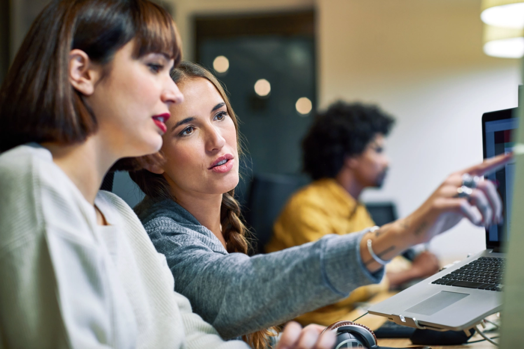 Two women collaborate at a laptop during a work meeting, pointing at the screen together.