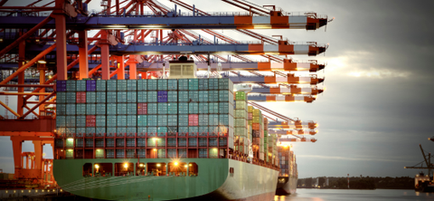 Photo of a green-hulled container ship berthed under a long row of red gantry cranes at dusk.