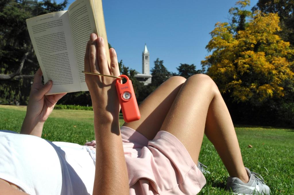 Person reading a book on a sunny lawn with a red Birdie safety alarm clipped to the cover