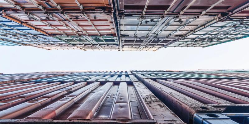 Upward view between two towering stacks of shipping containers converging toward a pale sky