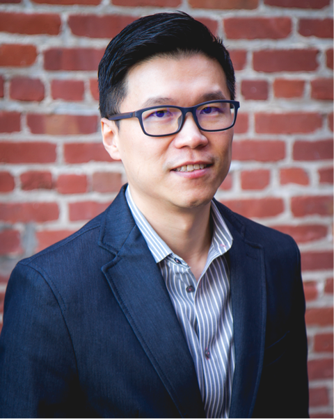Headshot of a man in a dark blazer and striped shirt against an exposed brick wall