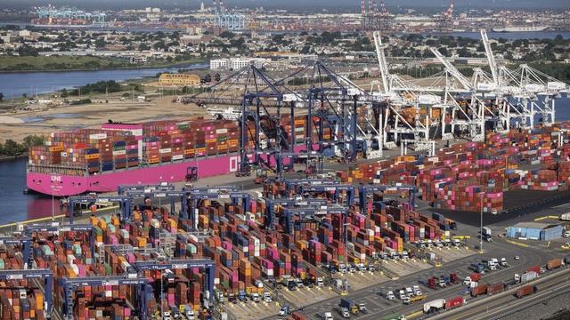 Aerial view of a busy container port with a pink-and-magenta cargo ship docked beside rows of stacked shipping containers and cranes