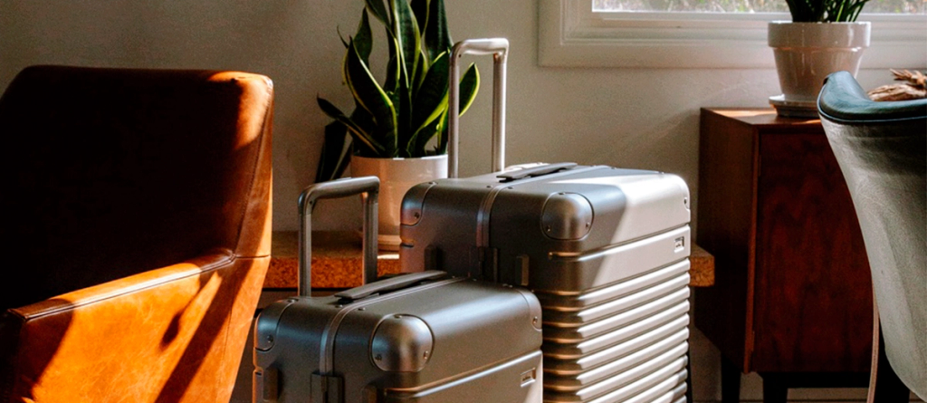Two silver hardshell suitcases standing on a floor beside a leather chair and houseplant