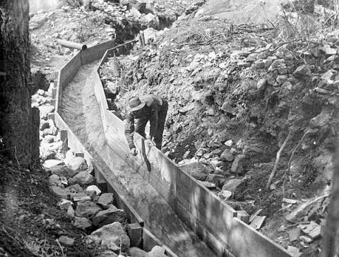 Grayscale historical photo of a man building a wooden flume sluice through a rocky dig site