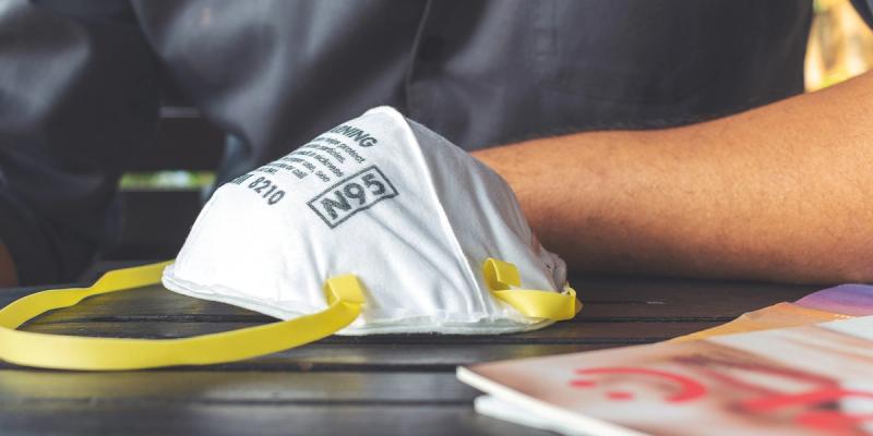 White N95 respirator mask labeled 8210 with yellow straps resting on a dark table by a person's arm