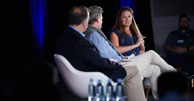 Three panelists seated on a conference stage, a woman gesturing as she speaks to two men