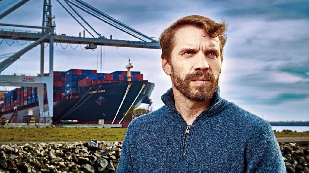 Bearded man in a blue quarter-zip sweater standing before a docked container ship and port cranes at the Port of Oakland