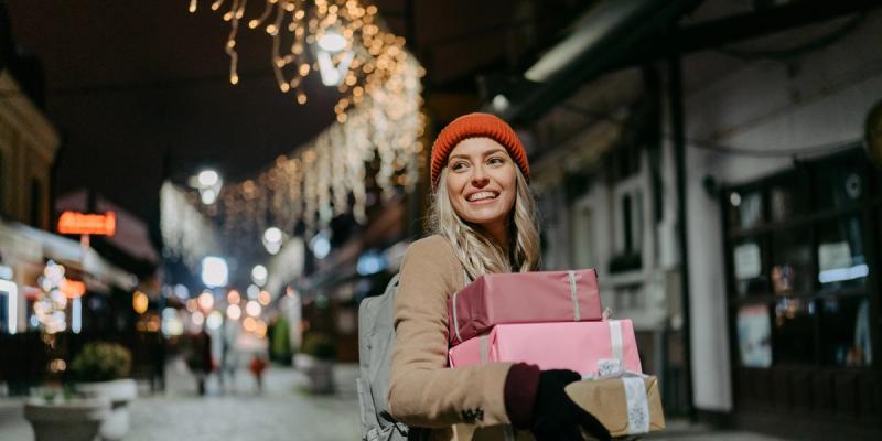Smiling woman in a red beanie carries wrapped gifts on a city street lit with string lights at night