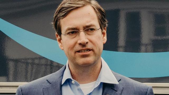 Man in a blue blazer and glasses speaking, standing in front of a dark backdrop with a teal swoosh