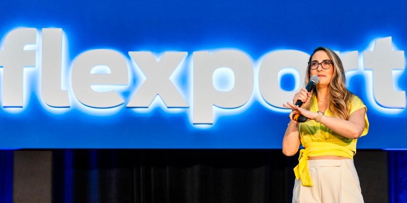 Woman in yellow blouse speaks into a microphone onstage before a glowing blue Flexport sign