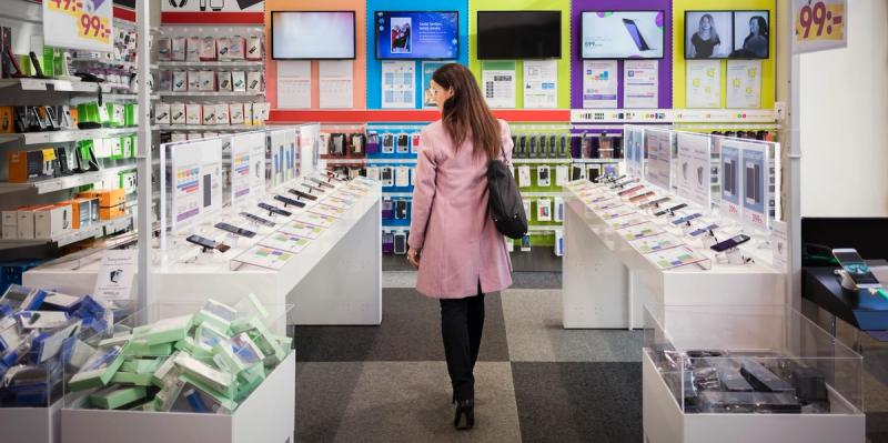 Woman in a pink coat walks through an electronics store lined with phone and case displays.