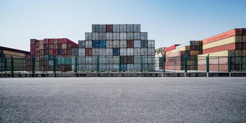 Stacked multicolored shipping containers behind a fence at a port terminal, empty paved lot in foreground