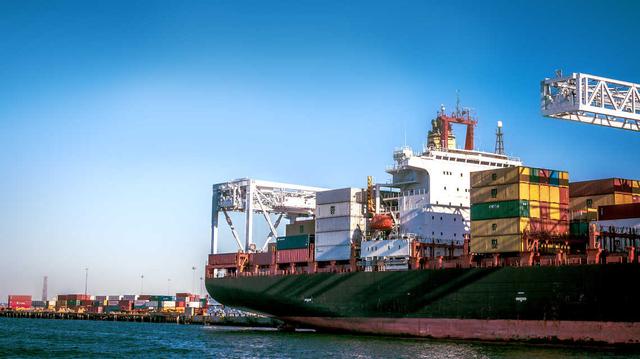 Container ship docked at a port with a loaded deck and a gantry crane reaching overhead under blue sky