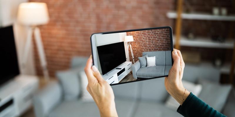 Hand holding a tablet that displays a living room with grey sofa and brick wall, overlaid on the same real room