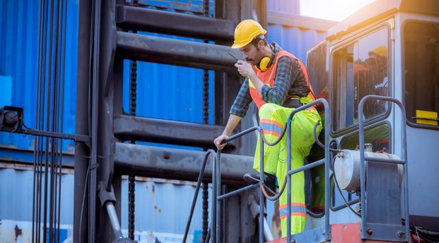 Dockworker in a hard hat and high-visibility gear speaks into a radio while climbing equipment beside stacked shipping containers