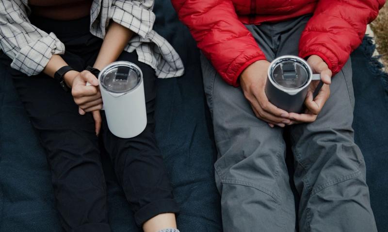 Two people outdoors on a blanket holding white and gray insulated travel mugs