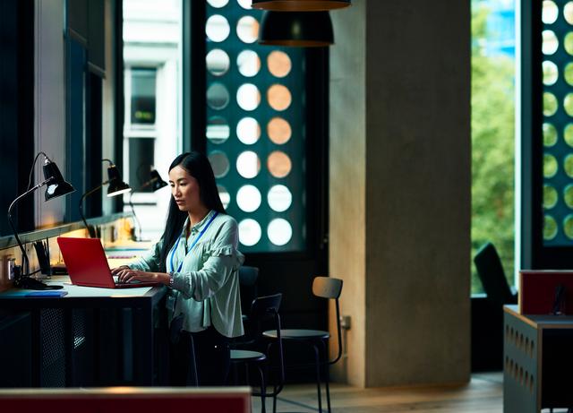 Woman with a lanyard works on a red laptop at a standing counter in a modern office with large windows