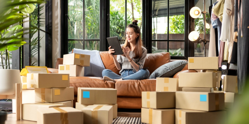 Woman sitting on a sofa using a tablet, surrounded by many cardboard parcels in a plant-filled home