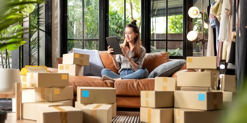 Woman sitting on a sofa using a tablet, surrounded by many cardboard parcels in a plant-filled home