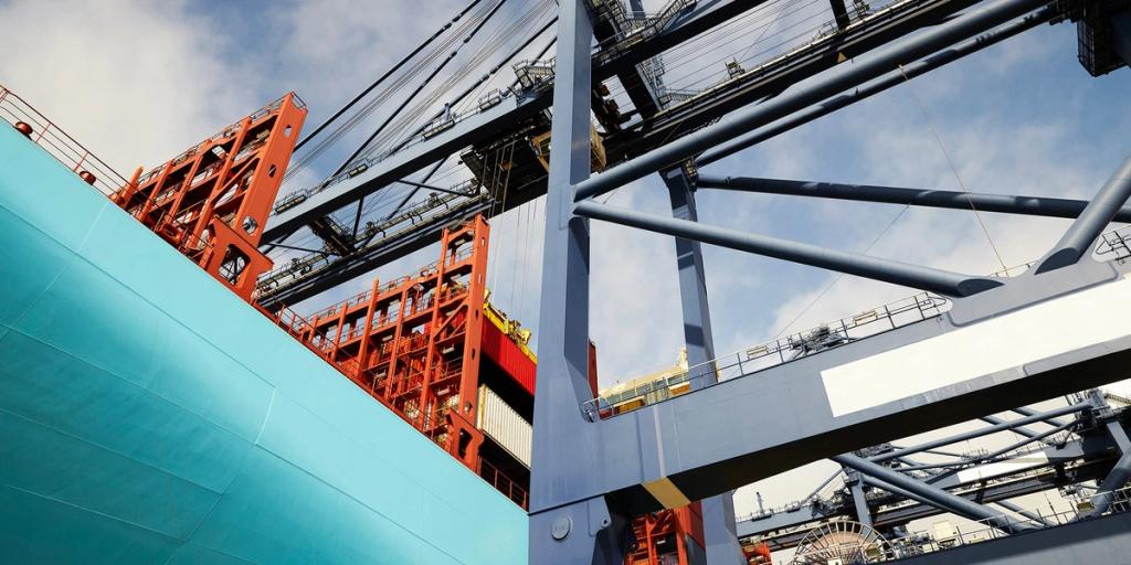 Low-angle view of a teal container ship hull beside red and grey port gantry cranes