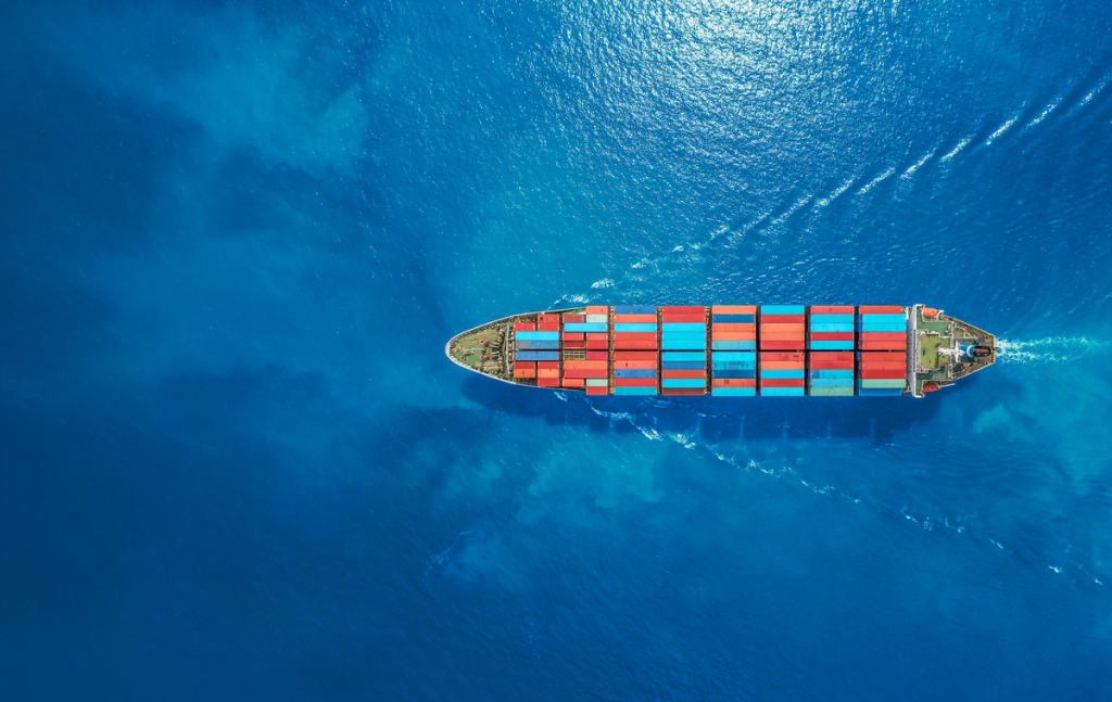 Cargo ship loaded with shipping containers traveling through open ocean, viewed from above.