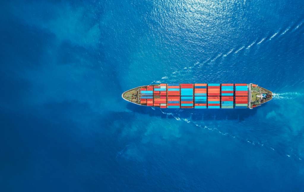 Cargo ship loaded with shipping containers traveling through open ocean, viewed from above.
