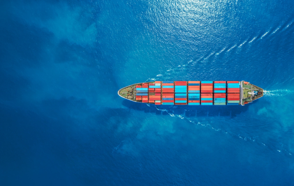 Cargo ship loaded with shipping containers traveling through open ocean, viewed from above.