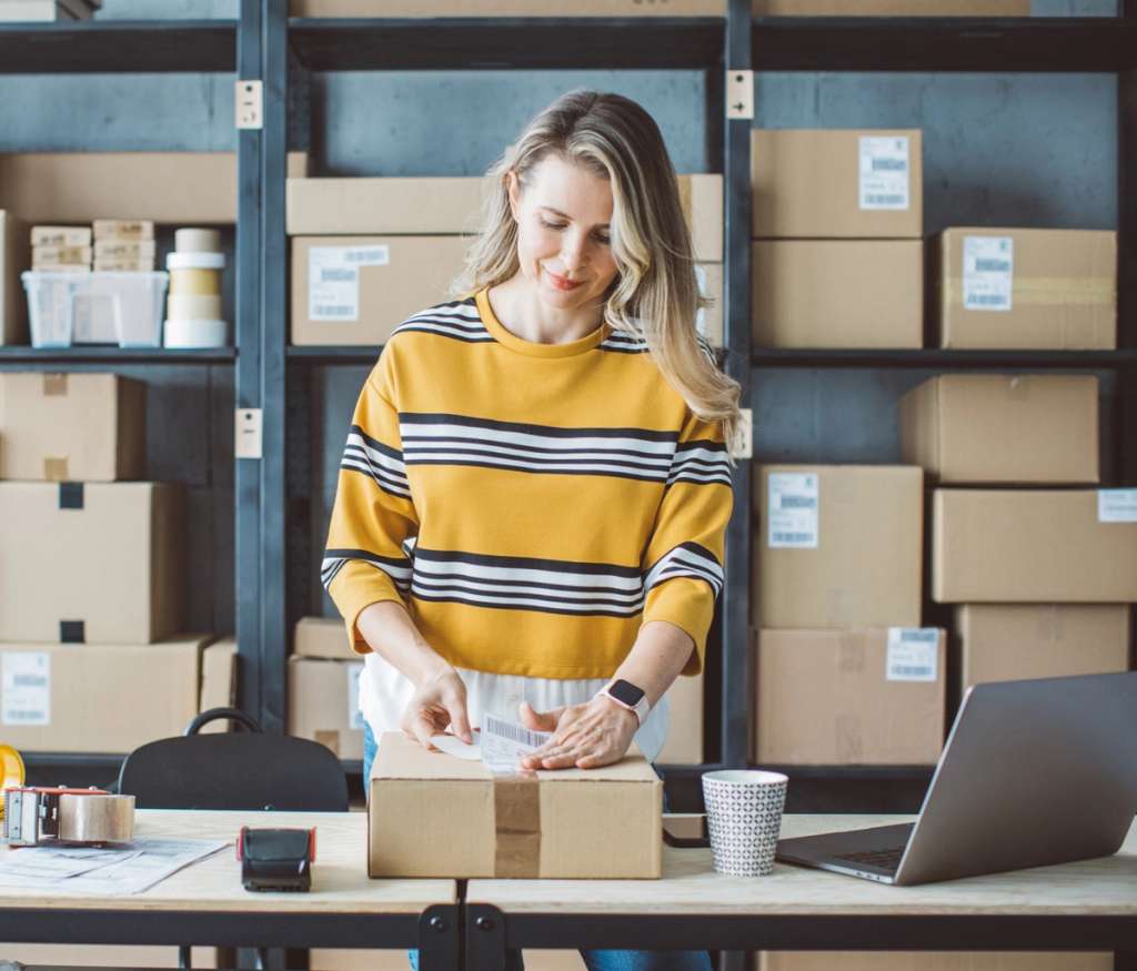 Woman in a striped sweater packing a cardboard parcel at a desk beside shelves of boxes and a laptop in a fulfillment workspace